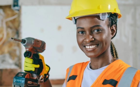 Confident female construction worker with hard hat and drill, smiling on site.